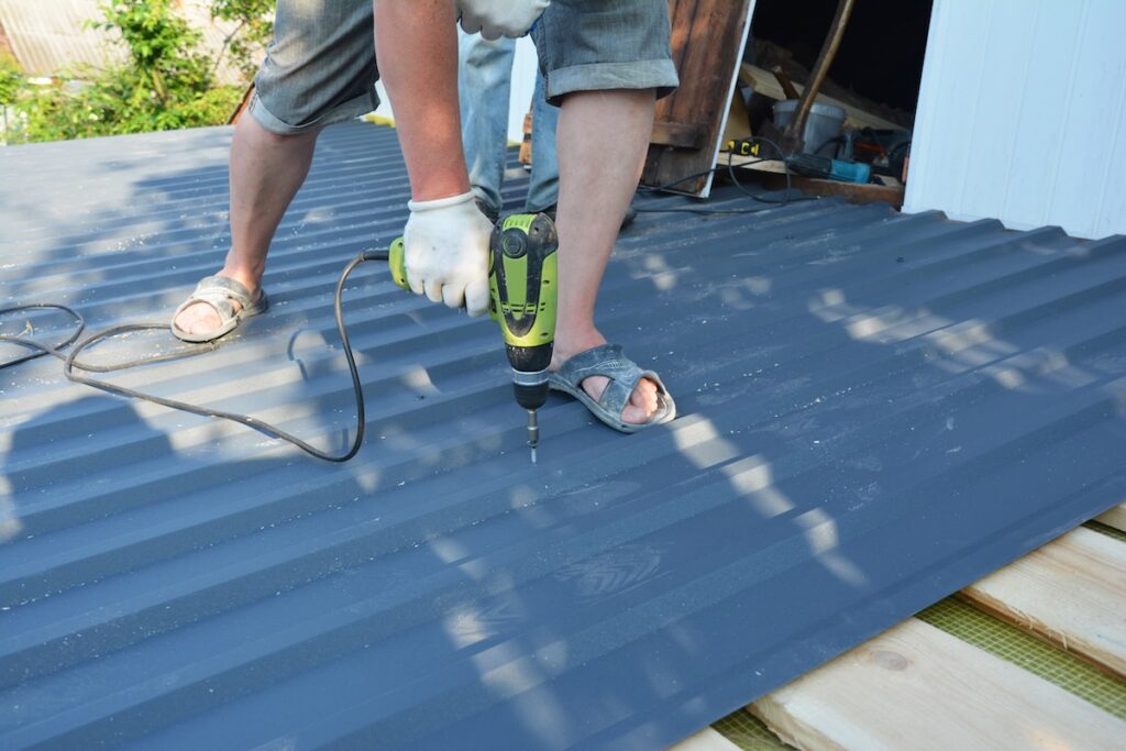 A roofing contractor is installing metal roof panels, screwing metal roofing sheet to the roof deck using an electric screwdriver.