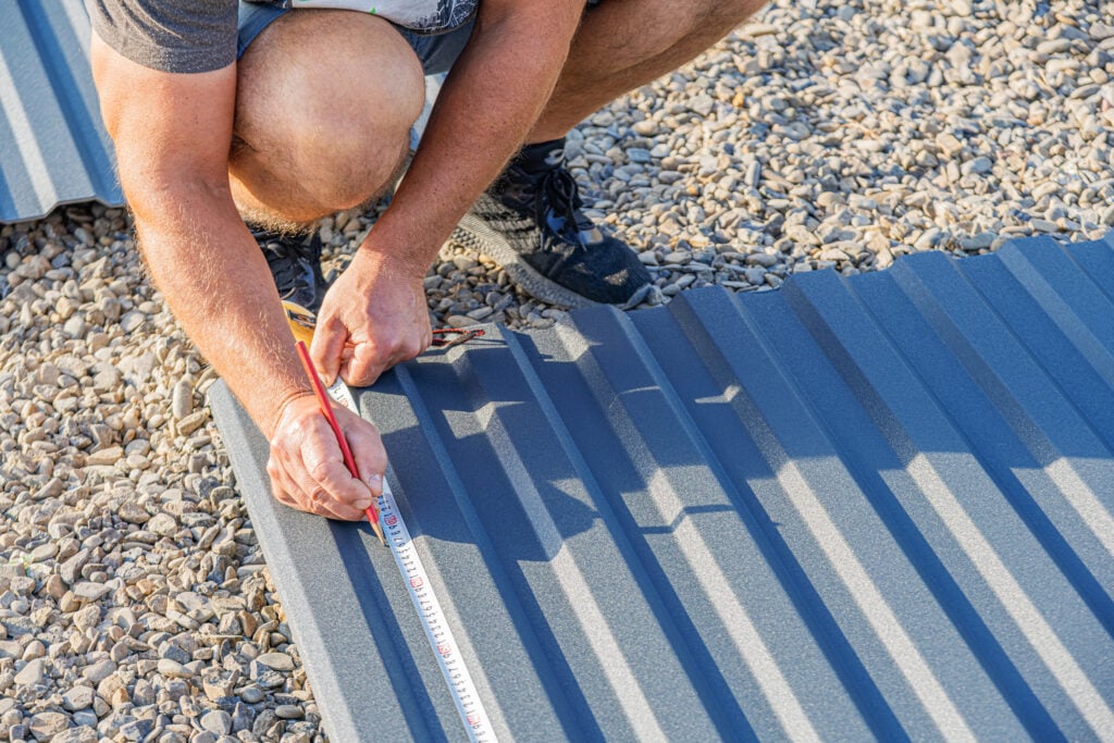 A man is measuring a corrugated metal sheet with a tape measure and pencil